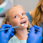 A young child sitting in a dental chair during a family dental care visit, as a dentist examines her teeth and an adult caregiver looks on.