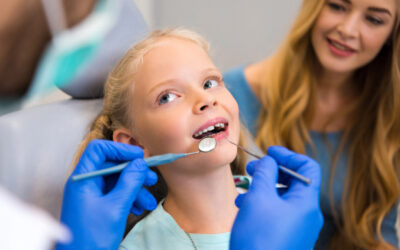 A young child sitting in a dental chair during a family dental care visit, as a dentist examines her teeth and an adult caregiver looks on.